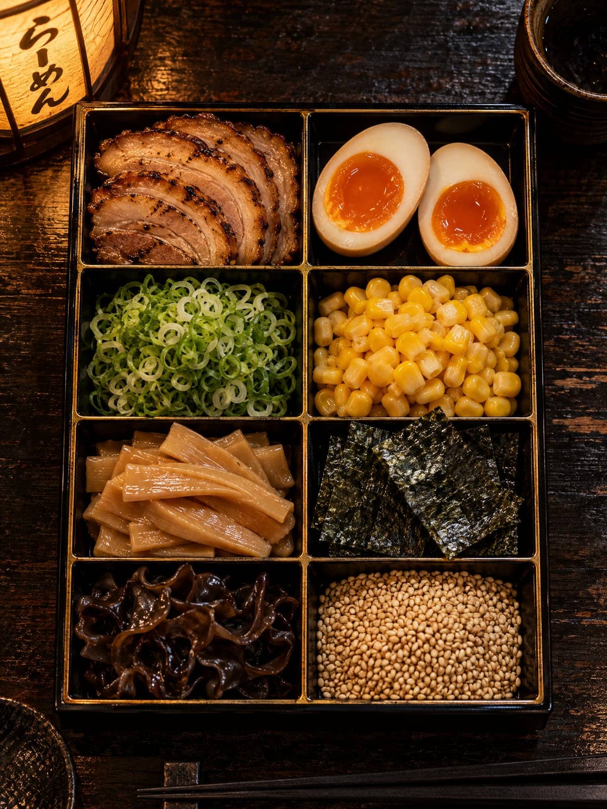 Overhead view of a black lacquer tray of ramen toppings: chashu, halved eggs, scallion, corn, bamboo shoots, nori, wood ear and sesame